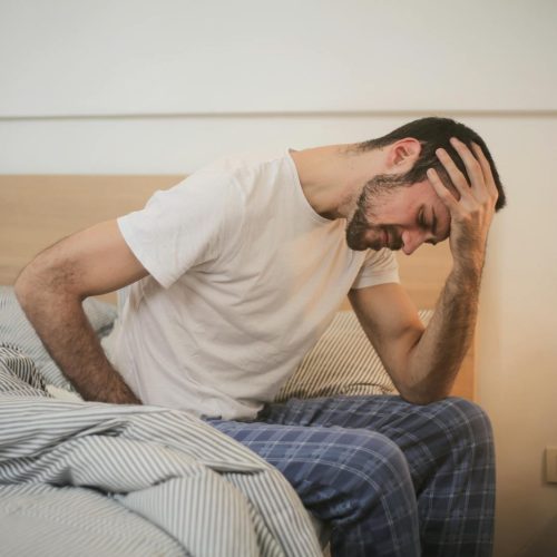 A young man in pajamas holding his head, sitting on a bed, appears to be experiencing a headache.